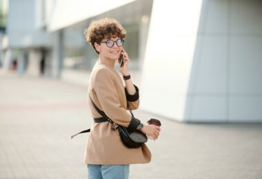 Contemporary young businesswoman with drink and handbag using smartphone